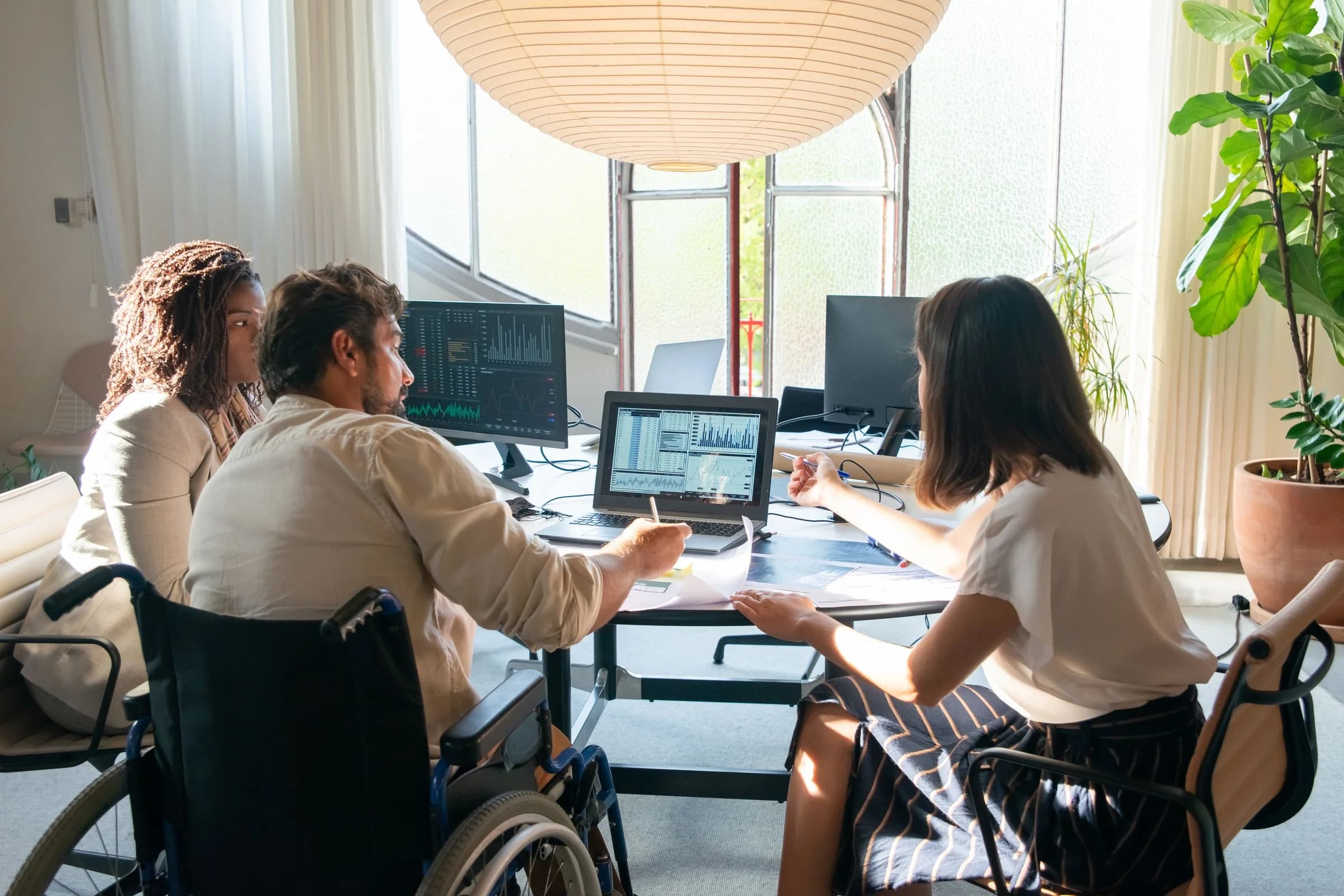 Three colleagues discussing charts on a computer beside a sunlit window