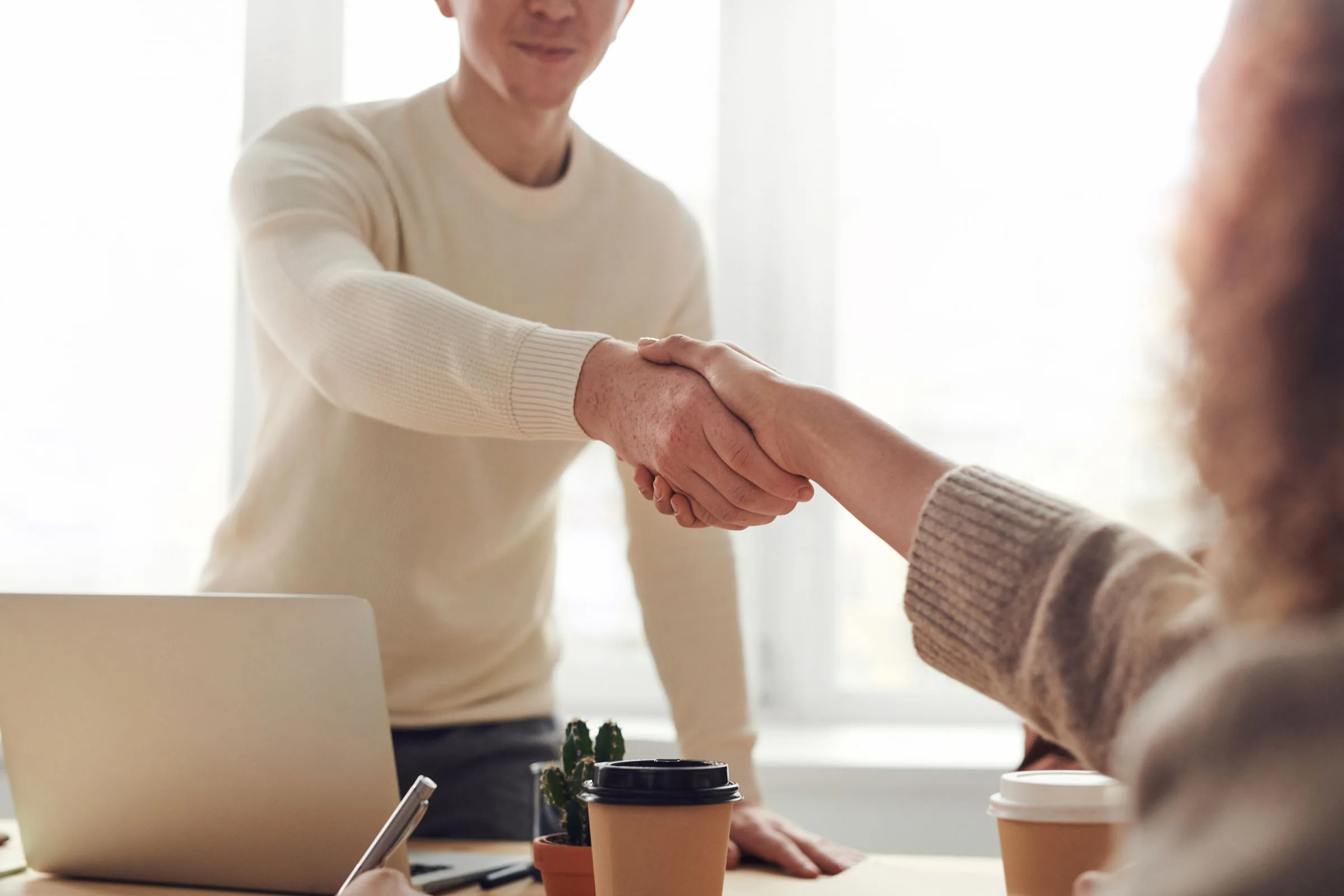 Two people shaking hands across a desk beside a laptop